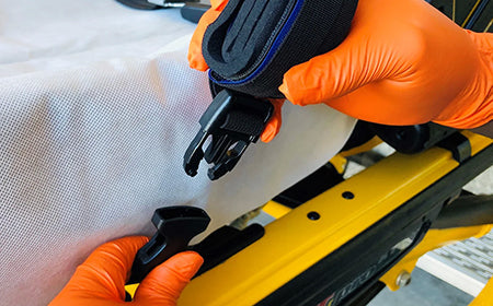 Gloved hands securing a black medical strap on a yellow EMS stretcher, Rowland Emergency equipment