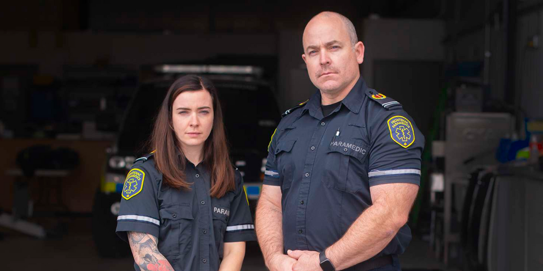 Two Frontenac County paramedics in uniform stand in front of an EMS vehicle in a garage
