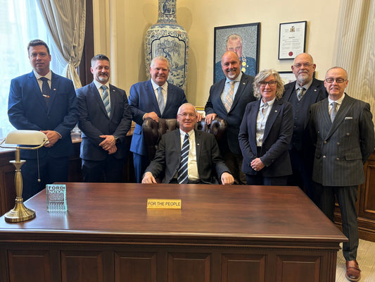 Group of Rowland Emergency team members in suits standing around a desk in a formal office with a 'For the People' nameplate, showcasing EMS custom solutions in Ontario.