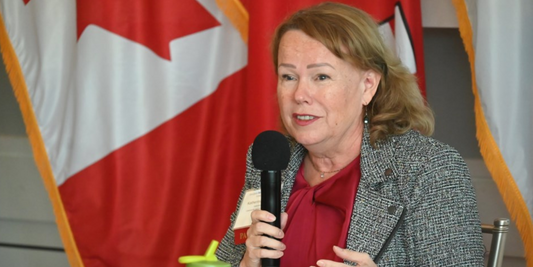Woman speaking into microphone at emergency preparedness event, Canadian flag in background
